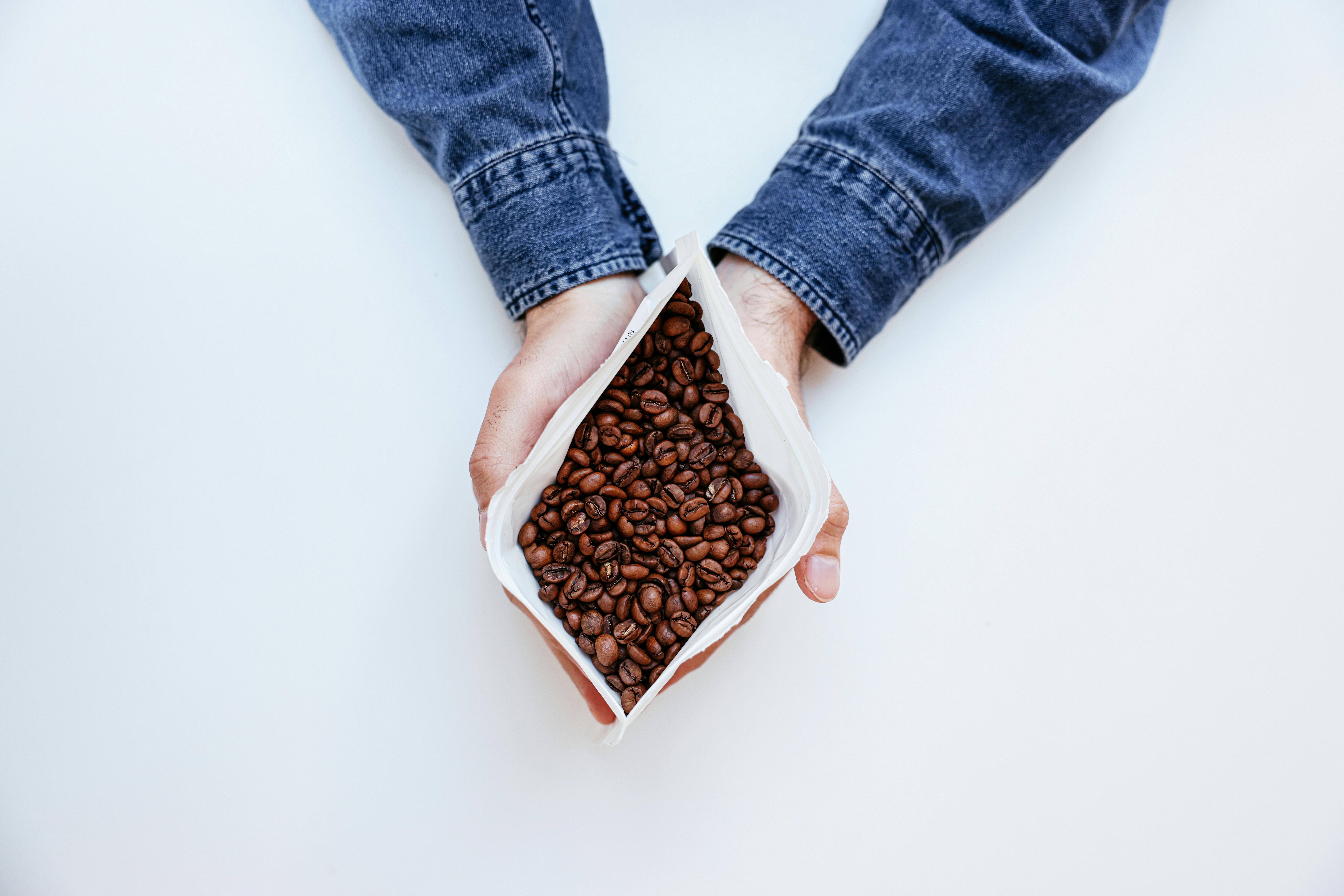 Hands holding an opened coffee bag filled with roasted beans, with the person wearing a denim shirt against a white background