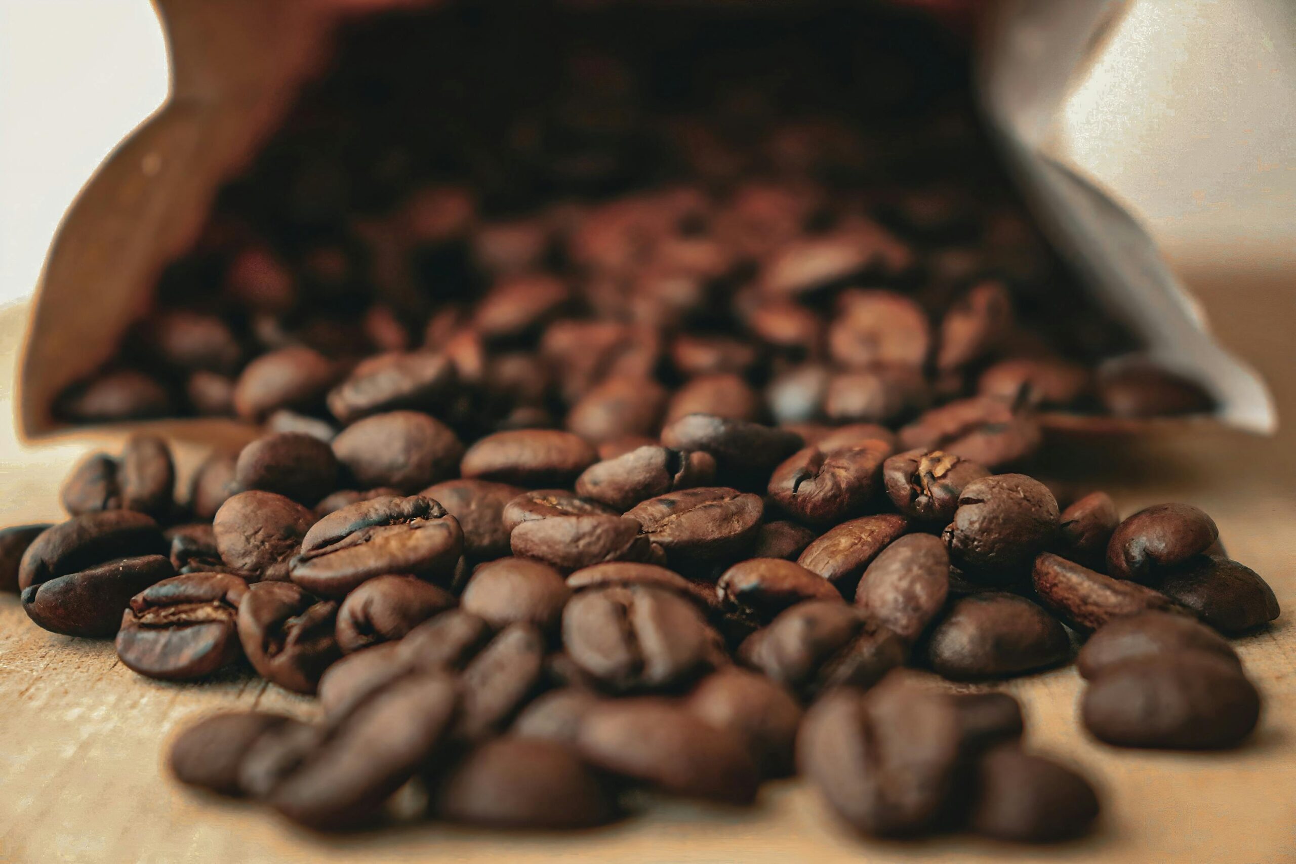 A close-up view of roasted coffee beans spilling out of an open brown paper bag onto a wooden surface. The beans are in sharp focus, while the background and the inside of the bag are softly blurred, emphasizing the rich texture and color of the coffee beans