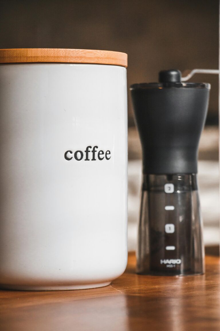 A ceramic container labeled "coffee" with a wooden lid, next to a manual coffee grinder. The container has a simple, minimalistic design, and the background is blurred, creating a cozy atmosphere