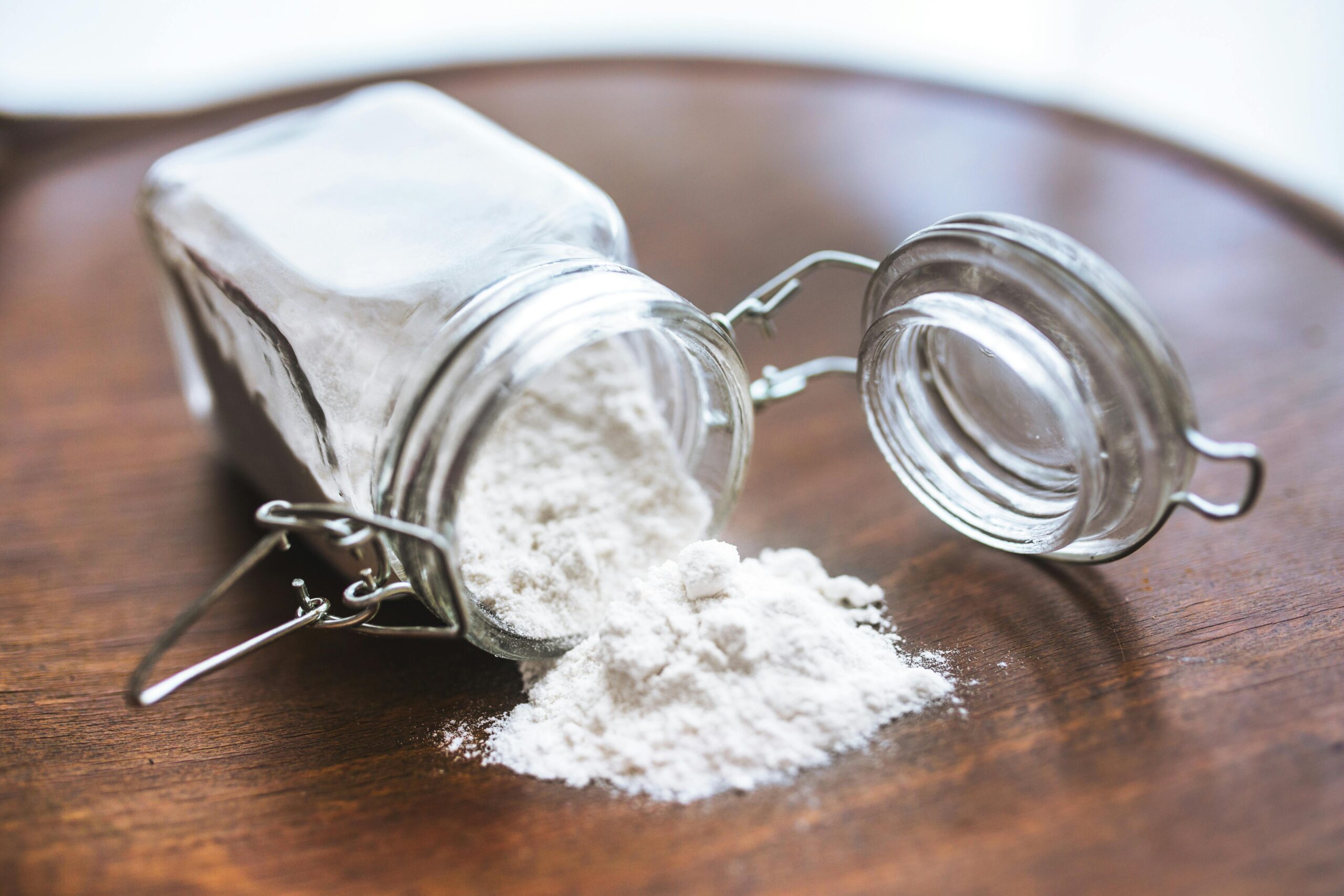A glass jar with an open lid lies on its side on a wooden surface, with a small mound of flour spilling out, creating a rustic, homely feel