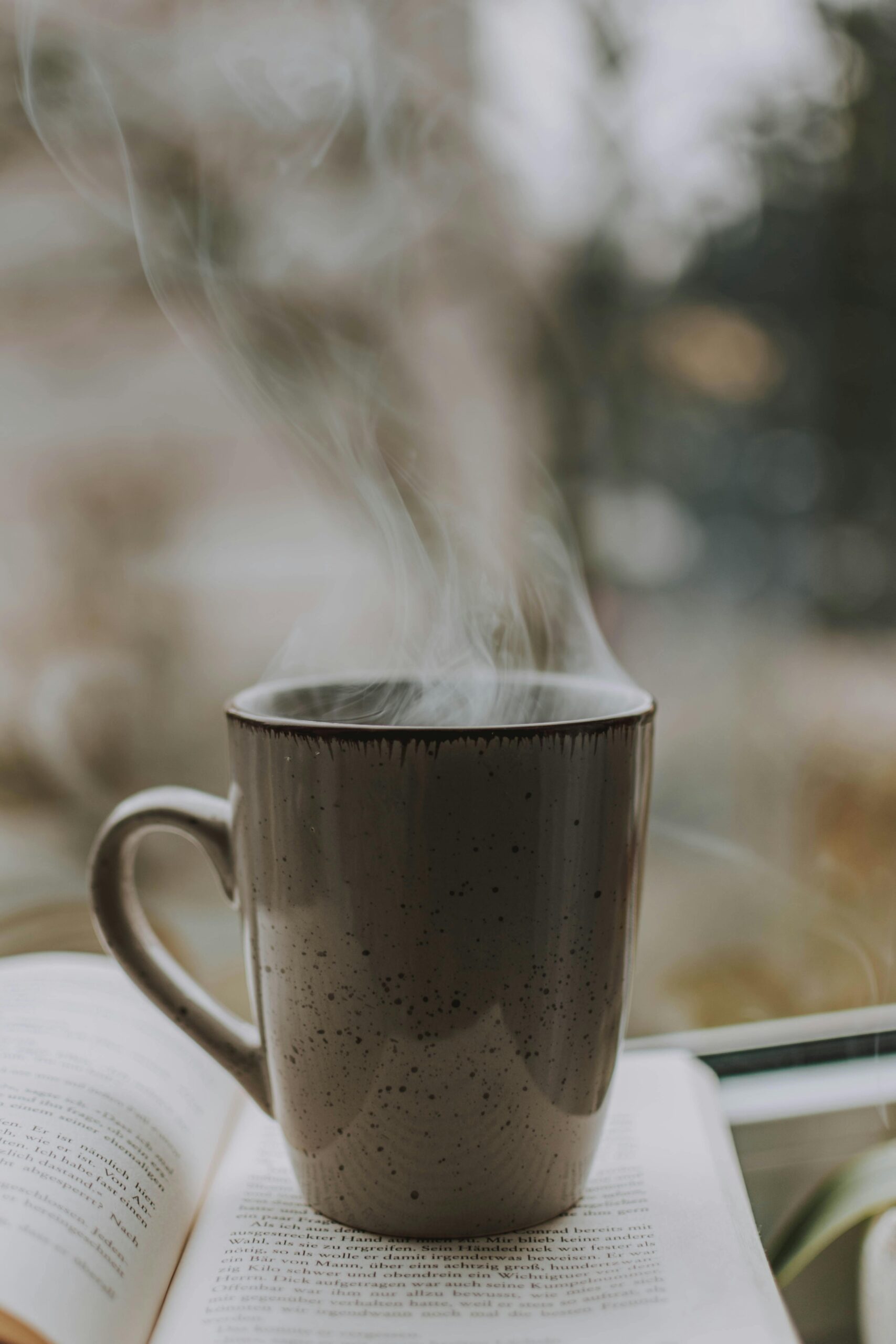 A steaming hot cup of coffee sits on top of an open book by a window, with soft light filtering through the background