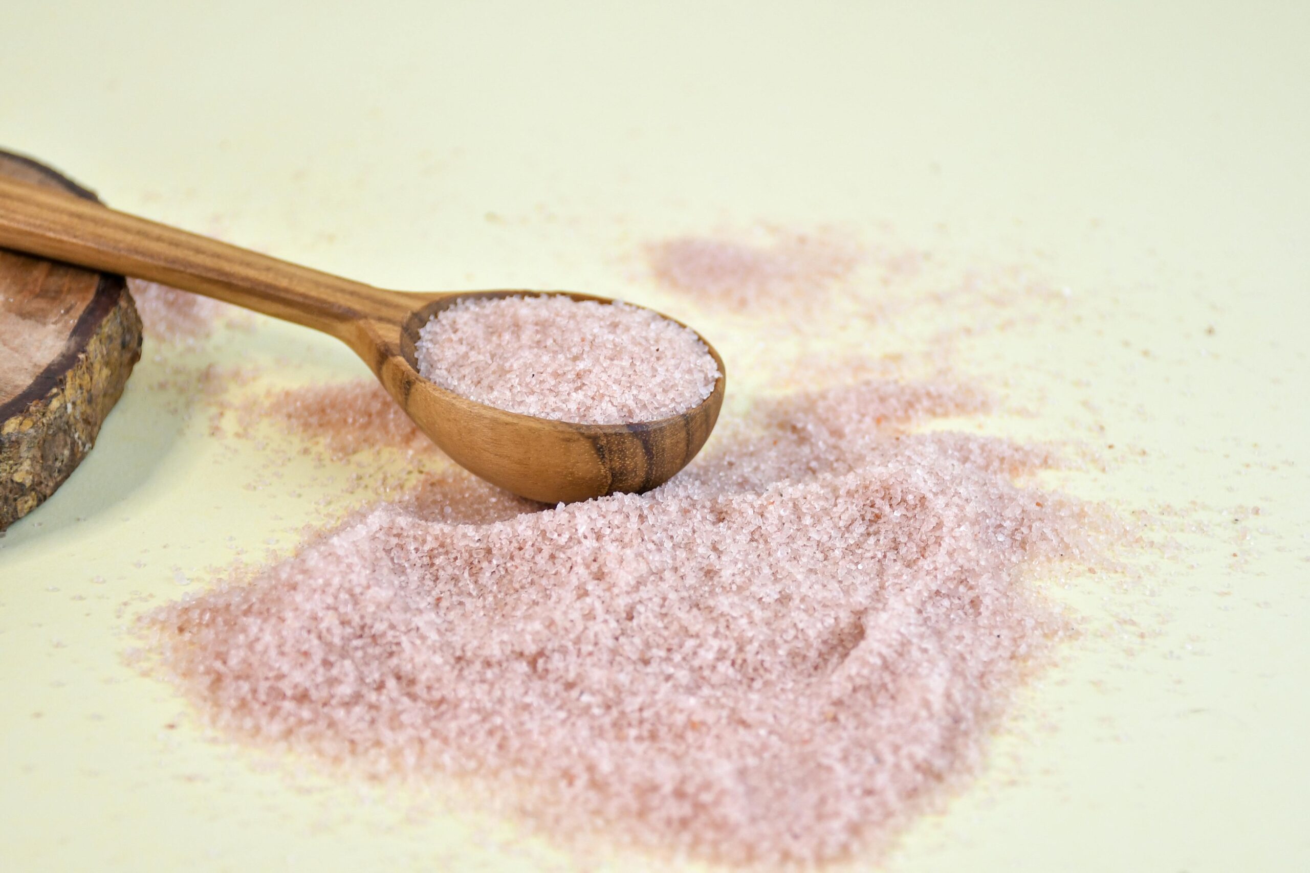 A wooden spoon filled with pink Himalayan salt lies on a pastel surface with scattered grains around it, giving a natural and earthy look