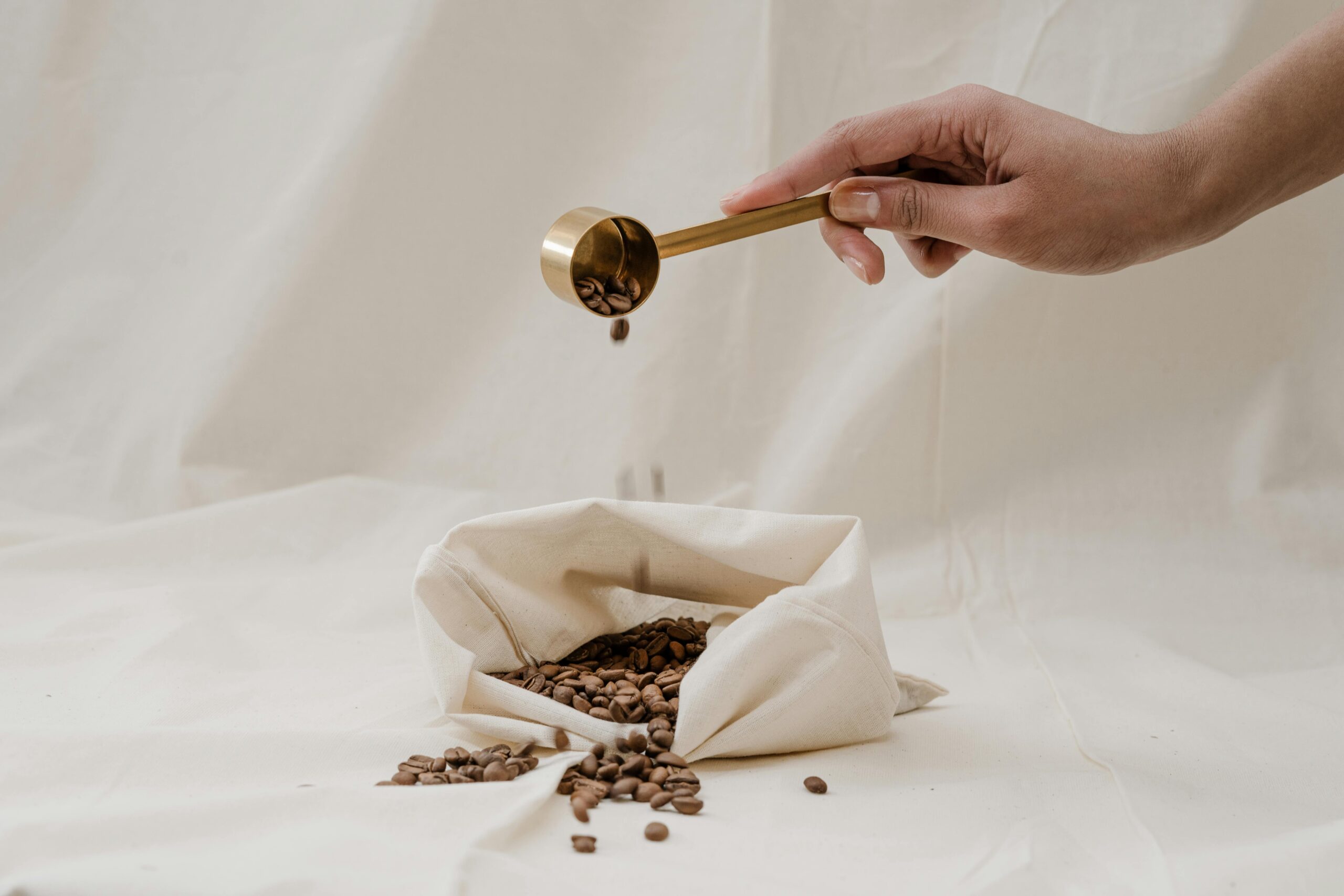 A hand holding a gold coffee scoop, gently pouring roasted coffee beans into a beige cloth bag, with scattered beans around on a neutral fabric background