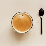 A top-down view of a glass cup filled with freshly brewed coffee, featuring a smooth crema layer on top. A black spoon is placed beside the cup on a clean, neutral-toned background